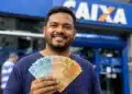 Homem brasileiro sorridente segurando cédulas de reais em frente à agência da Caixa Econômica Federal, ilustrando o saque de R$ 6.220 da Caixa.
