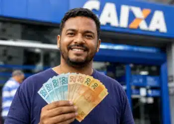 Homem brasileiro sorridente segurando cédulas de reais em frente à agência da Caixa Econômica Federal, ilustrando o saque de R$ 6.220 da Caixa.