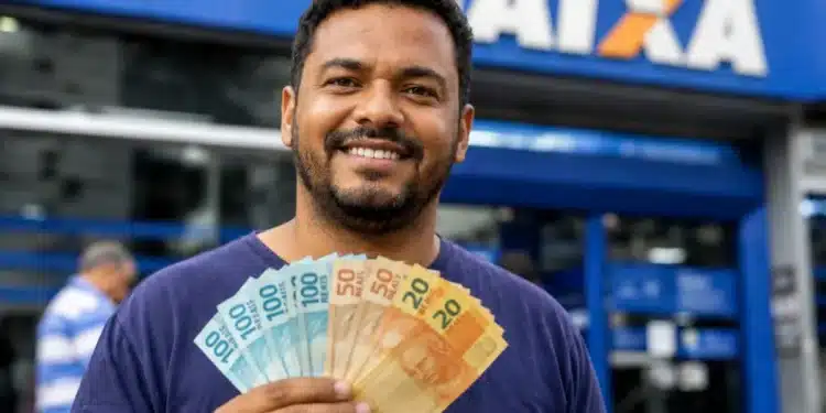 Homem brasileiro sorridente segurando cédulas de reais em frente à agência da Caixa Econômica Federal, ilustrando o saque de R$ 6.220 da Caixa.