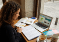 Mulher com cabelo cacheado estuda idiomas com livros, notebook aberto no Duolingo e café em mesa de cafeteria.