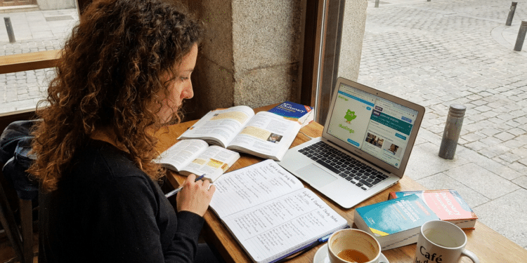 Mulher com cabelo cacheado estuda idiomas com livros, notebook aberto no Duolingo e café em mesa de cafeteria.
