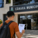 Candidato com mochila segura documento de concurso público em frente à Câmara Municipal.