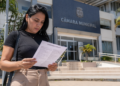 Mulher lê documento de concurso público em frente à Câmara Municipal, representando inscrições para vagas de nível fundamental e médio.