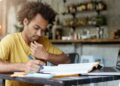 Jovem concentrado estuda apostila e faz anotações ao lado do notebook em preparação para concurso público.