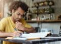 Jovem concentrado estuda apostila e faz anotações ao lado do notebook em preparação para concurso público.