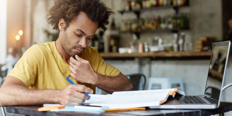 Jovem concentrado estuda apostila e faz anotações ao lado do notebook em preparação para concurso público.