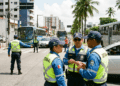 Quatro agentes de trânsito do DETRAN-AL com uniforme azul e colete amarelo, conversam em avenida de Maceió.