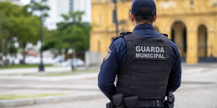 Guarda Municipal de costas com identificação no colete, representando concurso com 100 vagas e salário de até R$ 13.747,02.