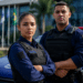 Dois guardas municipais brasileiros de braços cruzados frente a viatura azul, com bandeira do Brasil ao fundo.