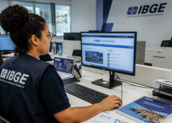 Funcionária do IBGE com colete azul-marinho usa computador acessando site do instituto em escritório com logo na parede.