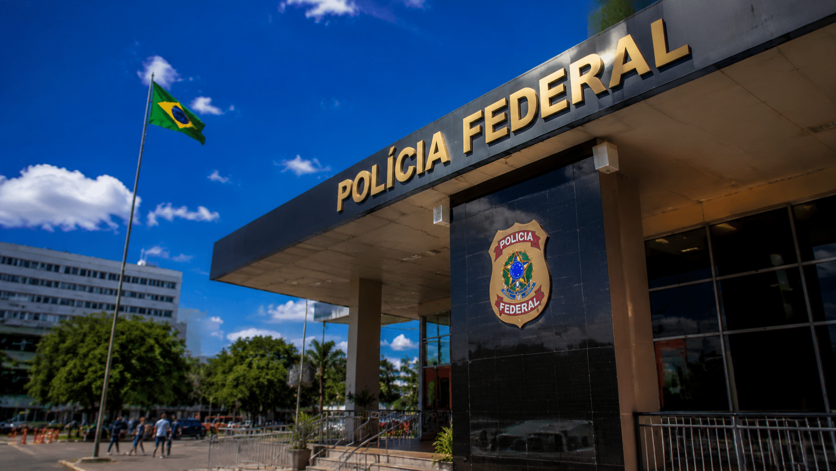 Fachada da Polícia Federal com brasão, bandeira do Brasil e céu azul ao fundo.