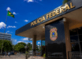 Fachada da Polícia Federal com brasão, bandeira do Brasil e céu azul ao fundo.