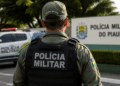 Policial da Polícia Militar do Piauí visto de costas em área externa, com viatura ao fundo.