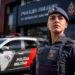 Policial feminina da PMESP usando boina e colete tático posa em frente à viatura e prédio da corporação.