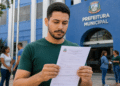 Homem segurando formulário na entrada da Prefeitura Municipal, pessoas conversando ao fundo, fachada azul.