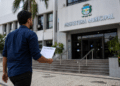 Homem caminhando em direção à prefeitura municipal segurando documento de concurso público.