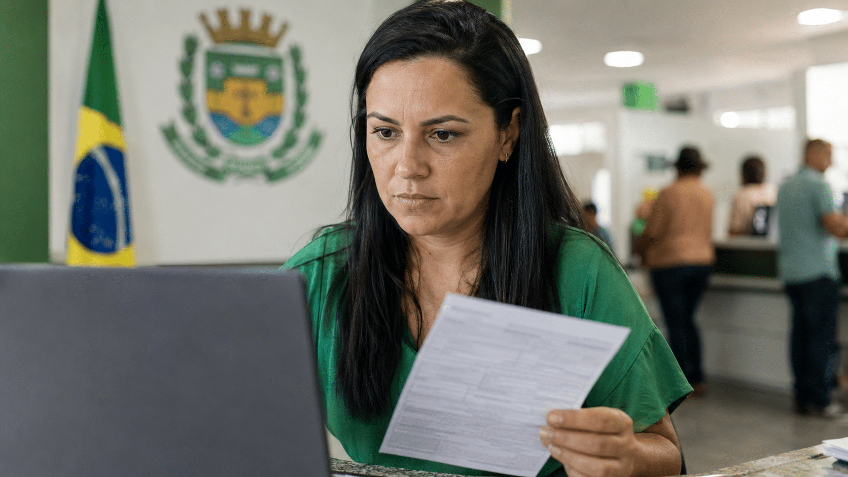 Mulher de cabelo escuro analisa formulário em frente a notebook em repartição pública com bandeira do Brasil e brasão oficial ao fundo.