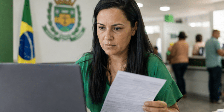 Mulher de cabelo escuro analisa formulário em frente a notebook em repartição pública com bandeira do Brasil e brasão oficial ao fundo.