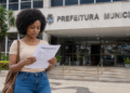 Mulher lê edital de concurso público em frente a uma prefeitura municipal, representando seleção com mais de 400 vagas.