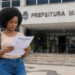 Mulher lê edital de concurso público em frente a uma prefeitura municipal, representando seleção com mais de 400 vagas.
