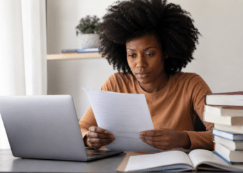Mulher estudando para concurso público em frente ao notebook, com documentos e livros sobre a mesa.