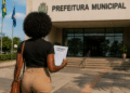 Mulher negra com cabelo afro caminhando em direção à sede da prefeitura municipal segurando edital de concurso público.