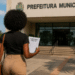 Mulher negra com cabelo afro caminhando em direção à sede da prefeitura municipal segurando edital de concurso público.
