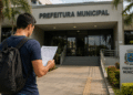 Homem caminhando em direção à Prefeitura Municipal enquanto lê documento de concurso público, representando edital com inscrições abertas.