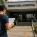 Homem caminhando em direção à Prefeitura Municipal enquanto lê documento de concurso público, representando edital com inscrições abertas.