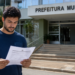 Homem lê documento de concurso público em frente a uma prefeitura municipal, representando a abertura de inscrições no RJ.
