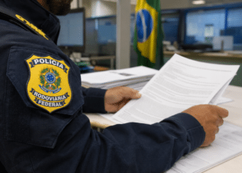Policial Rodoviário Federal uniformizado lê folhas de documentos em mesa de escritório com bandeira brasileira ao fundo.
