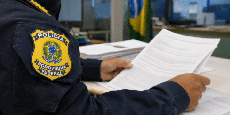 Policial Rodoviário Federal uniformizado lê folhas de documentos em mesa de escritório com bandeira brasileira ao fundo.