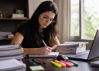 Jovem mulher de óculos estudando e escrevendo anotações à mesa cheia de livros e laptop.