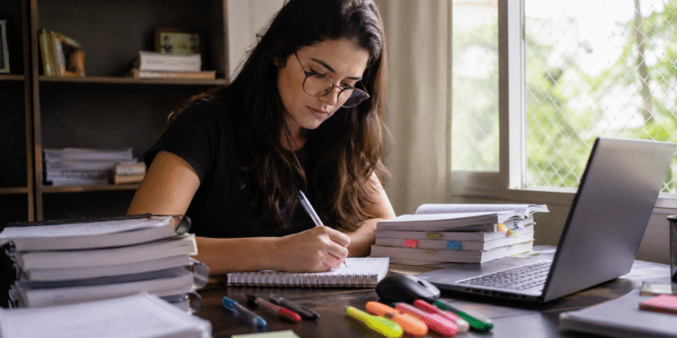 Jovem mulher de óculos estudando e escrevendo anotações à mesa cheia de livros e laptop.