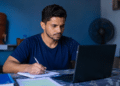 Homem escrevendo em caderno enquanto usa notebook em ambiente doméstico azul.