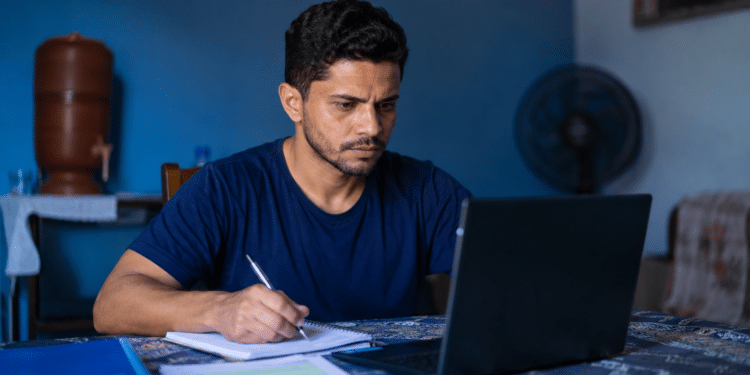Homem escrevendo em caderno enquanto usa notebook em ambiente doméstico azul.
