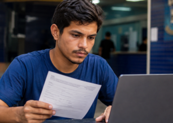 Jovem segura ficha de inscrição e consulta notebook em ambiente de órgão público para concurso da Prefeitura e da Câmara.