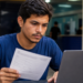 Jovem segura ficha de inscrição e consulta notebook em ambiente de órgão público para concurso da Prefeitura e da Câmara.
