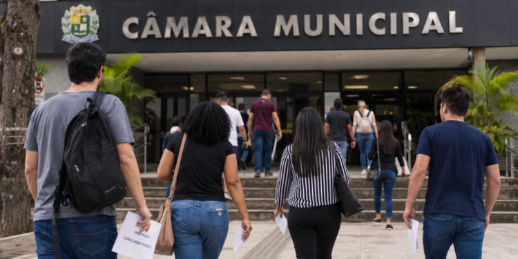 Pessoas caminham em direção à Câmara Municipal segurando documentos de concurso público.