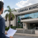 Pessoa em frente à Câmara Municipal segurando edital de concurso público.