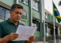 Homem de meia-idade lê papel com expressão séria diante de prédio verde com bandeira do Brasil hasteada.