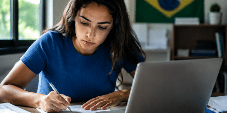 Mulher de camiseta azul escreve em documento enquanto consulta os concursos das Câmaras Municipais de Mara Rosa (GO) e Ipaporanga (CE) no laptop prateado, bandeira do Brasil ao fundo.