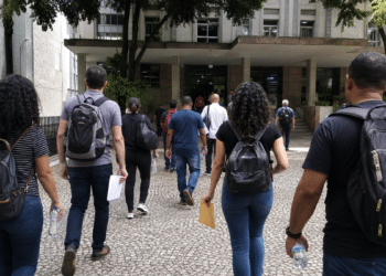 Pessoas com mochilas e documentos caminham em direção a prédio público para participar de seleções municipais.