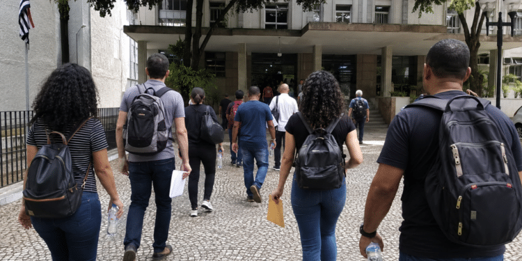 Pessoas com mochilas e documentos caminham em direção a prédio público para participar de seleções municipais.