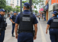 Quatro agentes da Guarda Municipal patrulham a calçada de um centro urbano com lojas e pedestres ao fundo.