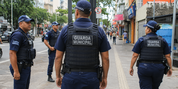 Quatro agentes da Guarda Municipal patrulham a calçada de um centro urbano com lojas e pedestres ao fundo.
