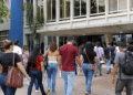 Pessoas caminhando em direção a prédio público com mochilas, bolsas, papéis e garrafas de água, representando candidatos de concursos em São Paulo.