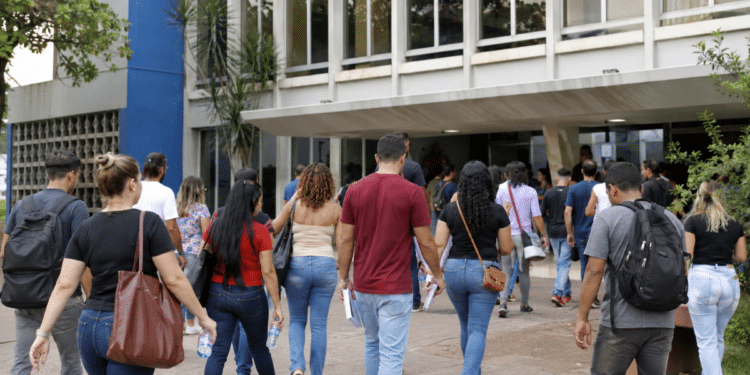 Pessoas caminhando em direção a prédio público com mochilas, bolsas, papéis e garrafas de água, representando candidatos de concursos em São Paulo.