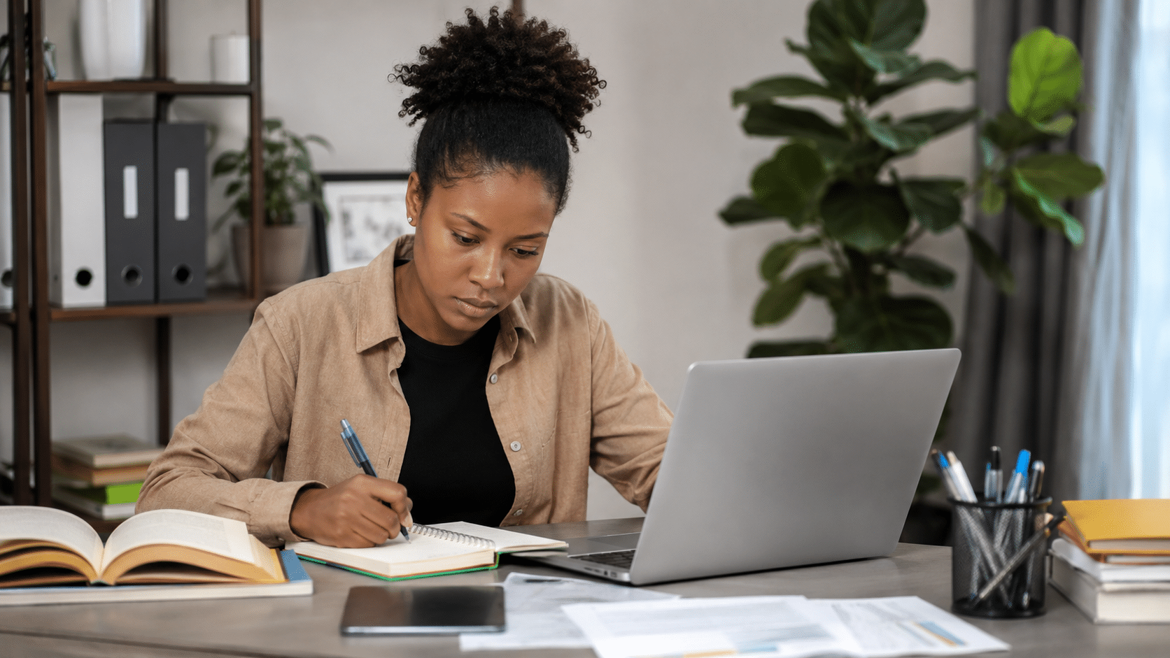 Mulher escrevendo em caderno ao lado de notebook e livros, ambiente de escritório caseiro.