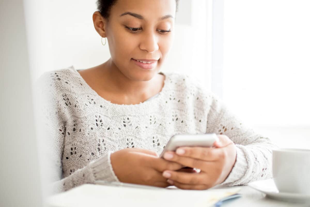Mulher sorri enquanto consulta o celular sentada à mesa, com xícara ao lado